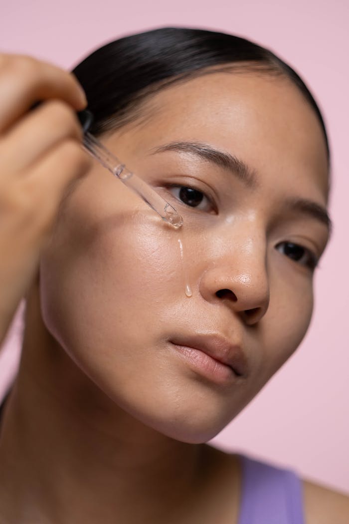 Close-up of a woman applying skincare serum with a dropper against a pink background.